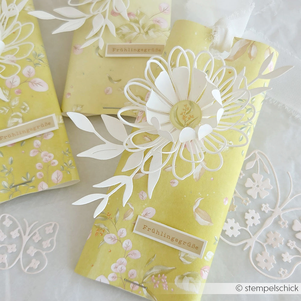 Drei gelbe Geschenkverpackungen mit Blumenmotiven und weißen Papierblumen, gestaltet mit der Motivstanze von Alexandra Renke und Anhängern mit der Aufschrift „Frühlingsgrüße“, dekorativ arrangiert.