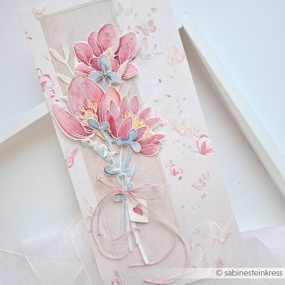 Handgefertigte Karte mit rosa Papierblumen, pastellfarbenen Blättern und einer Schleife auf einem hellrosa Hintergrund mit Schmetterlingsmuster – gefertigt mit der Motivstanze von Alexandra Renke und hochwertigen Designpapieren für ein elegantes Finish.