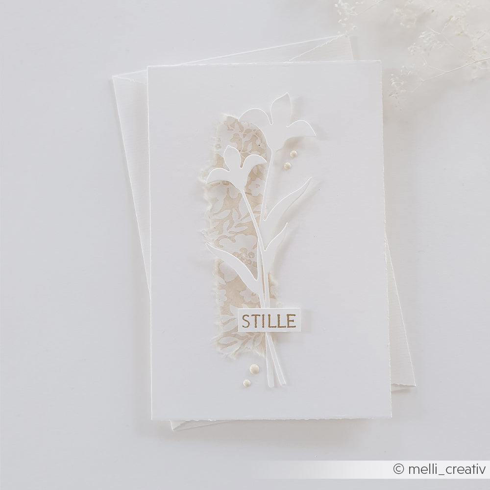 Weiße Karte mit zarten floralen Ausschnitten, die mit den „Motivstanze Blütenkomposition“-Stanzen von Alexandra Renke gestaltet wurden, mit einem Spitzenmuster und dem Schriftzug „STILLE“ auf weißem Hintergrund.