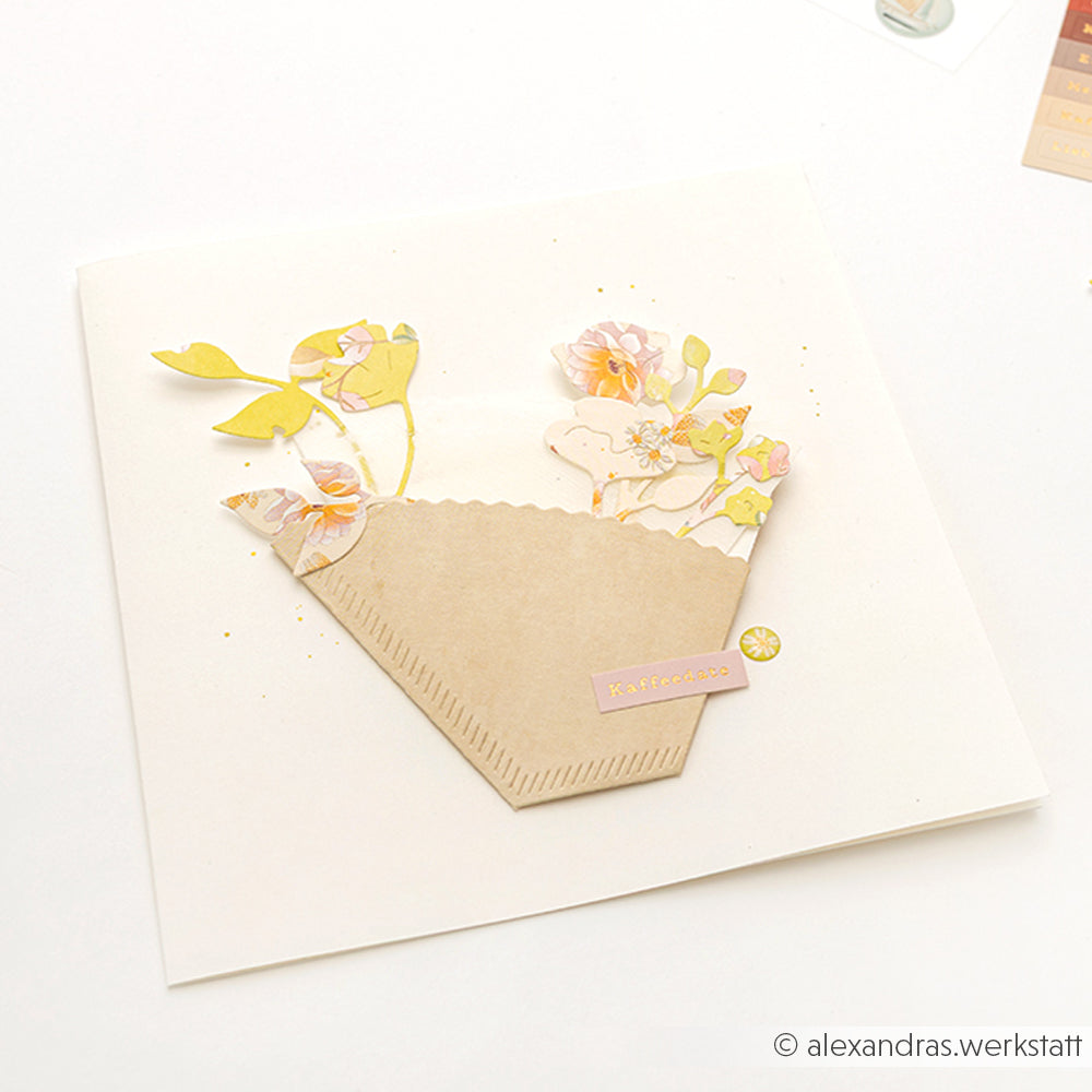 Handgefertigte Karte von Alexandra Renke mit pastellfarbenen Blumen in einem Kaffeefilter aus Papier auf weißem Hintergrund. Die eleganten Blüten sind mit der Motivstanze Gartengrüße gestaltet und verleihen ihr zusätzlichen Charme.