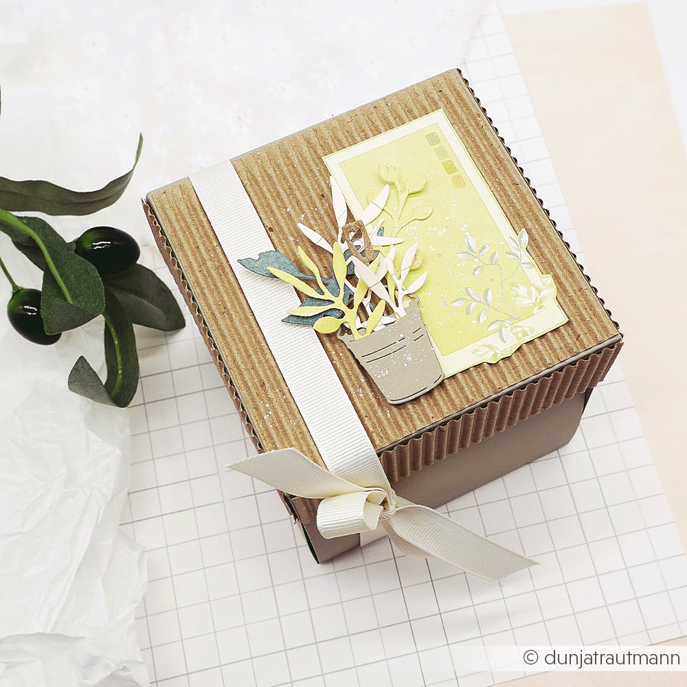 Quadratische Geschenkbox mit einer weißen Schleife, verziert mit einer Papierpflanze, die mit den „Gartengrüße“-Stanzformen von Alexandra Renke hergestellt wurde, sowie einem gelben Anhänger, auf kariertem Papier platziert. Ideal für kreative Projekte mit den Stanzsystemen von Alexandra Renke.