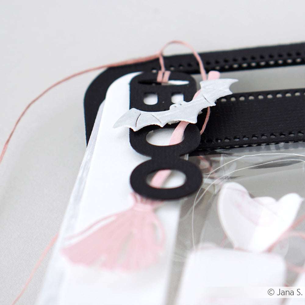 Nahaufnahme eines schwarzen BOO-Anhängers mit weißer Fledermaus und rosa Schnur auf einem Bastelprojekt, hergestellt mit Alexandra Renke Designpapieren und der Motivstanze Fledermausschwarm aus ihren beliebten Stanzsystemen.