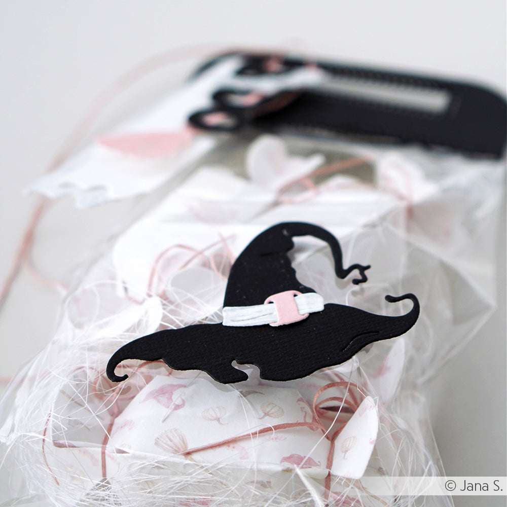 Geschenkverpackung aus weißem Netzstoff mit schwarzem Hexenhut, rosa Akzenten und einem Anhänger. Verarbeitet mit Alexandra Renke Designpapieren und Motivstanze Hexenzubehör Stanzsysteme für extra magische Details.