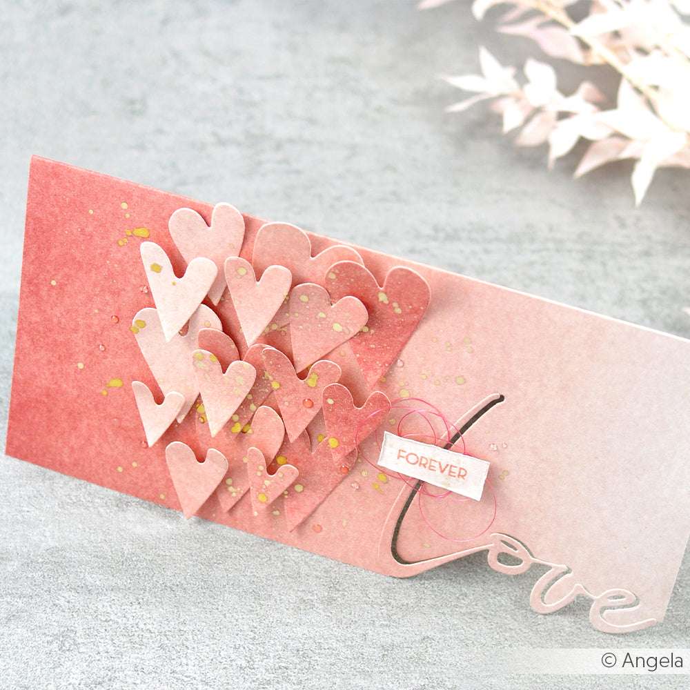 Handgefertigte rosafarbene Karte von Alexandra Renke, mit übereinanderliegenden Designpapieren und zarten Herzstanzungen, mit dem Wort "Liebe" und einem kleinen "Für immer"-Anhänger.