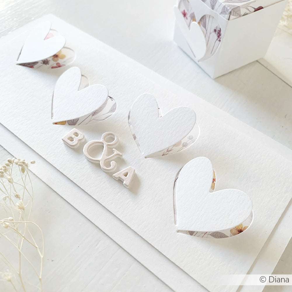 Weiße Hochzeitskarte mit Pop-up-Herzen aus der Alexandra Renke Motivstanze Herz mit dem Text "B & A". Präsentiert auf einer hellen Oberfläche mit floralen Akzenten für einen eleganten Touch.