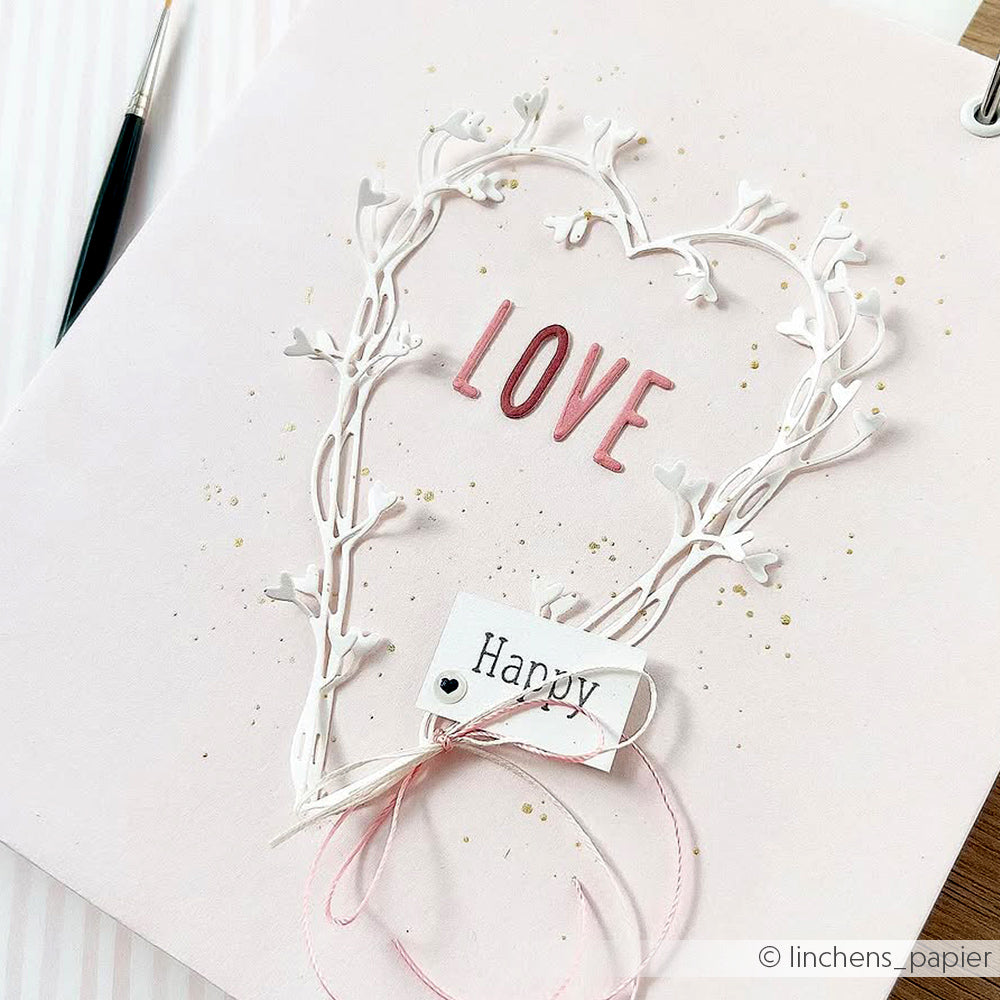 Eine handgefertigte Karte mit der Motivstanze Ranken-Herz von Alexandra Renke in Herzform, dem Schriftzug LOVE" in Rosa und einem mit rosa Schnur gebundenen Anhänger Happy".