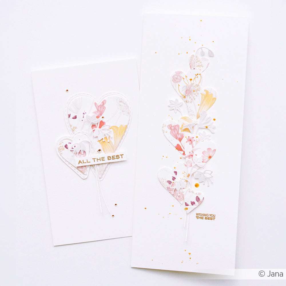 Zwei weiße Grußkarten mit floralen Herzmotiven und goldenem Text von Alexandra Renke auf weißem Hintergrund, hergestellt mit dem Stanzen für Designpapier und dem Motivstanze Herz-Tag Set für präzise Stanzdetails.