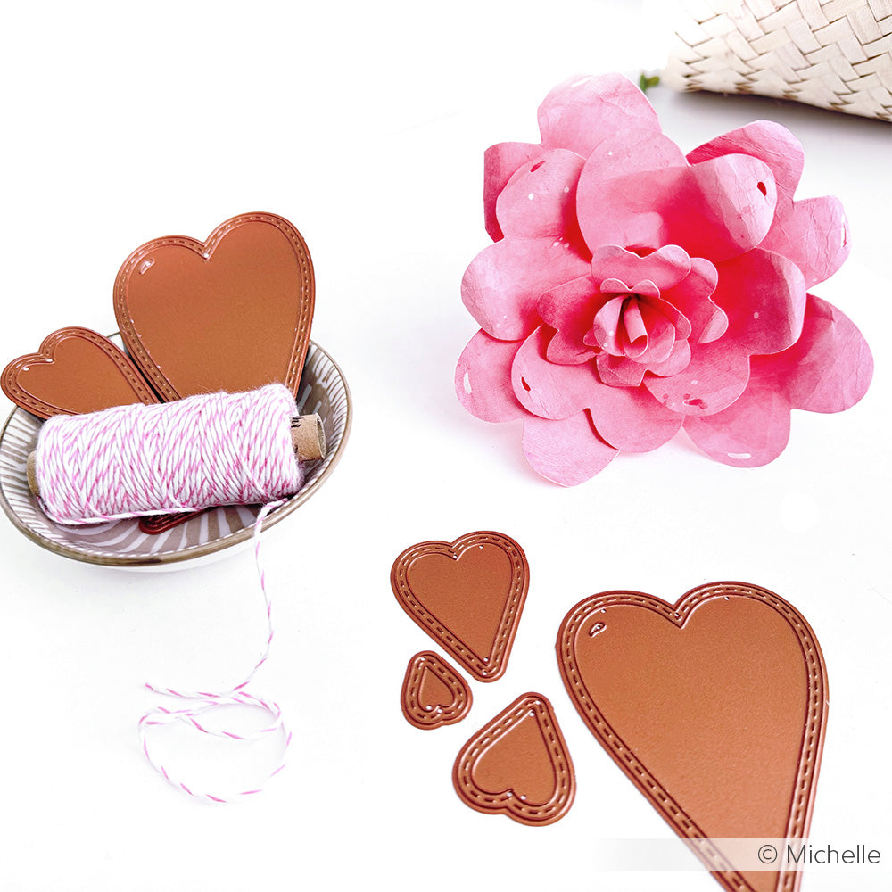 Auf einer weißen Fläche liegen herzförmige Bastelutensilien wie das Alexandra Renke Motivstanze Herz-Tag Set, rosa Schnur und eine rosa Papierblume aus Alexandra Renke Stanzen für Designpapier.