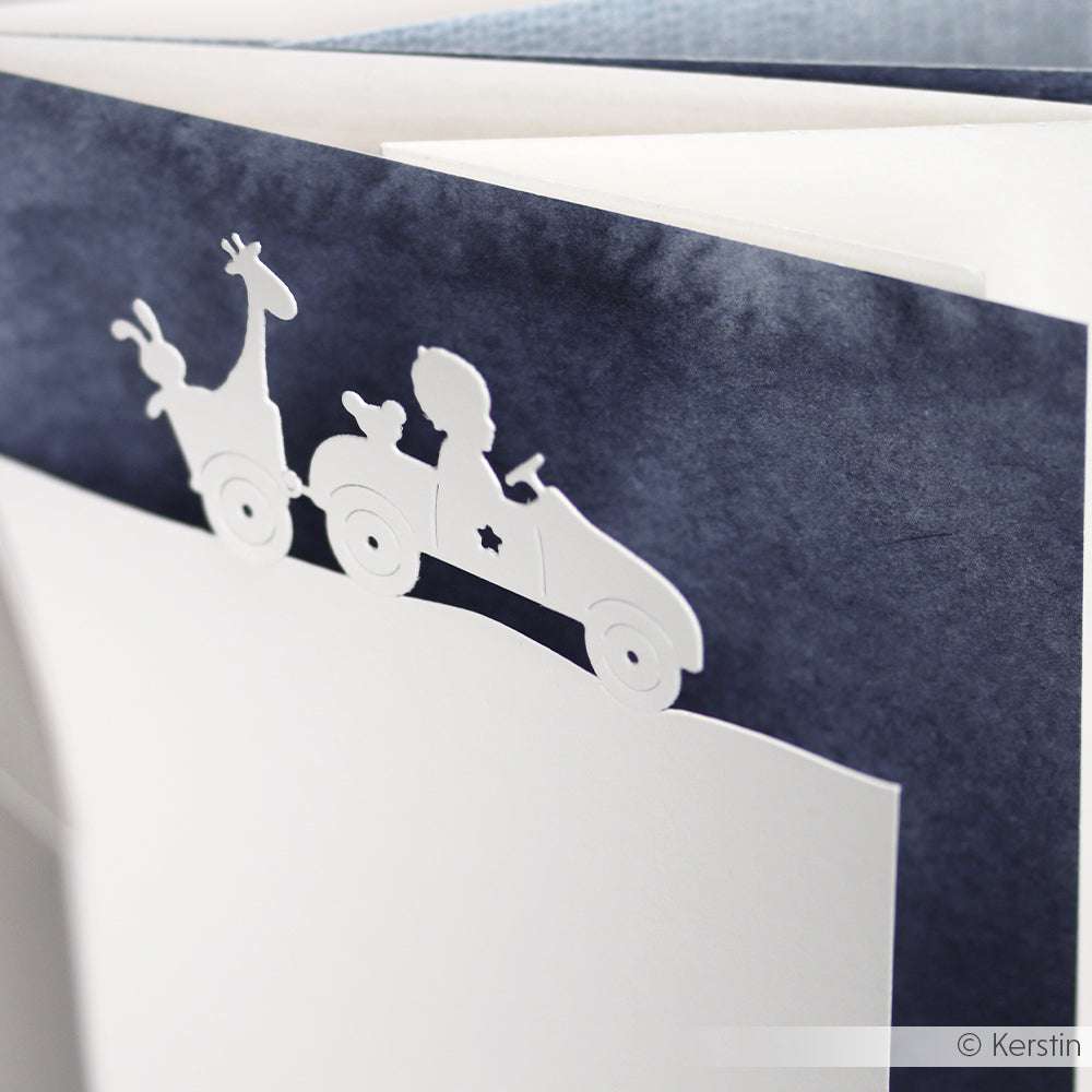 Weißer Papierausschnitt eines Kindes, das mit einer Giraffe in einem Auto fährt, hergestellt mit Alexandra Renke's DieCut Systeme Motivstanze Auto mit Anhänger, auf dunkelblauem Hintergrund.