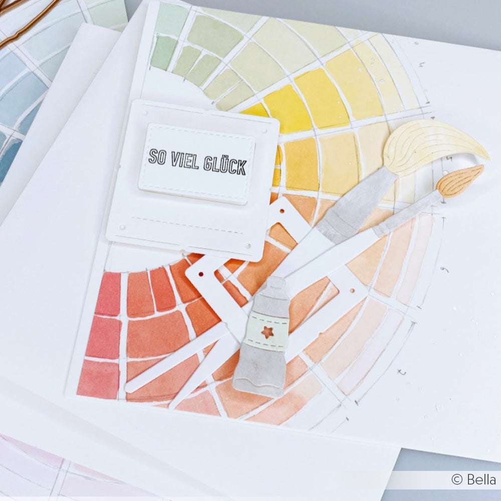 Ein Aquarell-Farbkreis mit Pinsel, Farbtube und einer SO VIEL GLÜCK-Notiz auf weißem Papier - die perfekte Inspiration, um die DieCut Systeme oder das Motivstanze Farbtuben Set von Alexandra Renke in Ihren Designpapier-Projekten einzusetzen.