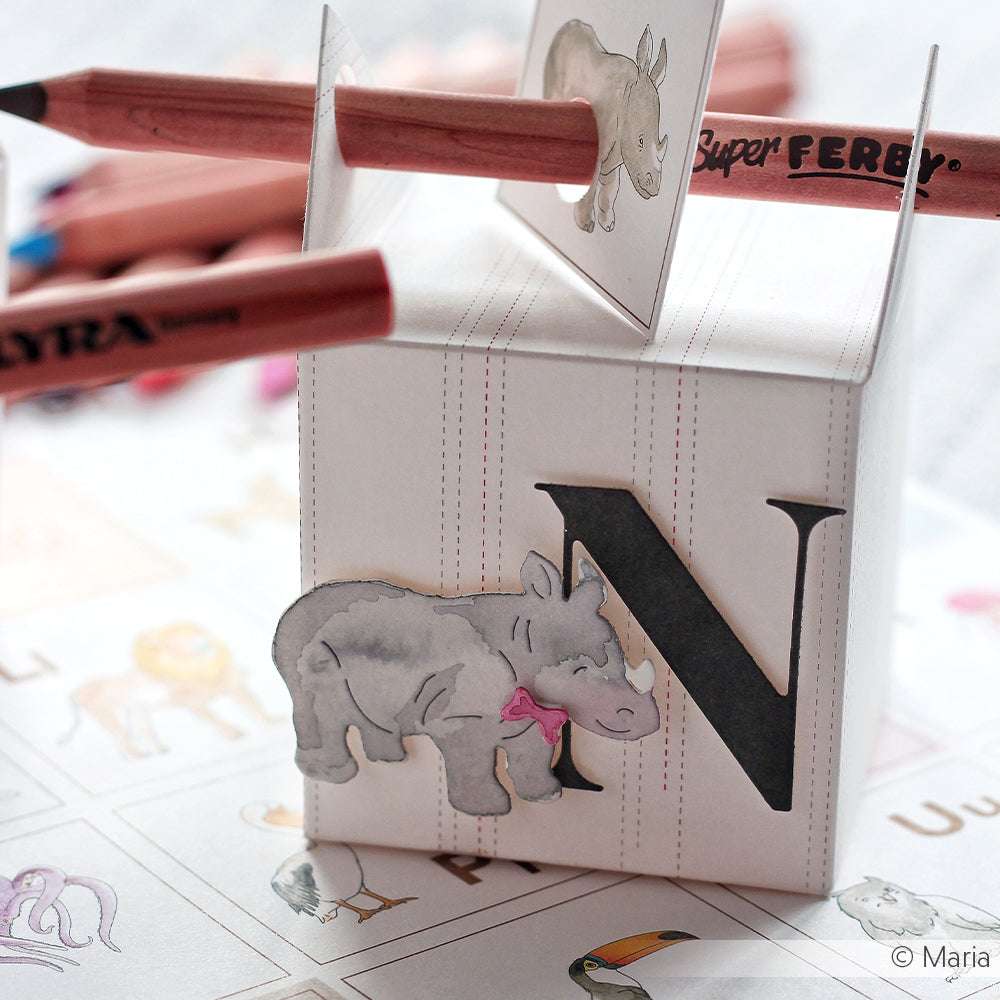 Ein Papierwürfel mit dem Buchstaben N und einer Nashorn-Illustration, hergestellt mit der Motivstanze Tierparade von Alexandra Renke, wird auf einem Arbeitsblatt mit Buntstiften durchstochen.