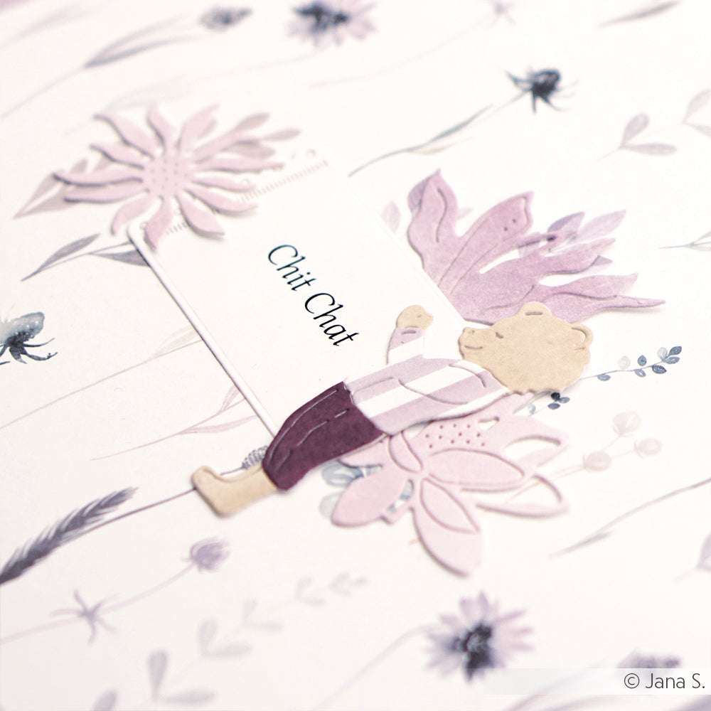 Ein Papierbär, der mit den Stanzformen von Alexandra Renke hergestellt wurde, trägt lila Kleidung und hält eine Karte mit der Aufschrift "Chit Chat" auf einem blumig gemusterten Hintergrund.