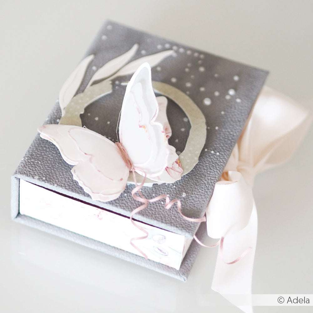 Graue Geschenkbox von Alexandra Renke mit einem rosa Schmetterling aus der Motivstanze Schichtschmetterling, weißem Band und zart strukturiertem Designpapier auf dem Deckel.