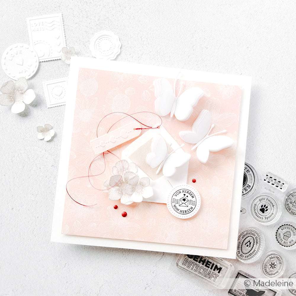 Quadratische Grußkarte mit weißen Schmetterlingen aus der Alexandra Renke Motivstanze Schichtschmetterling sowie Blumen, Stempeln und roten Details auf einem rosa gemusterten Hintergrund - perfekt für alle Papierbastelfans.