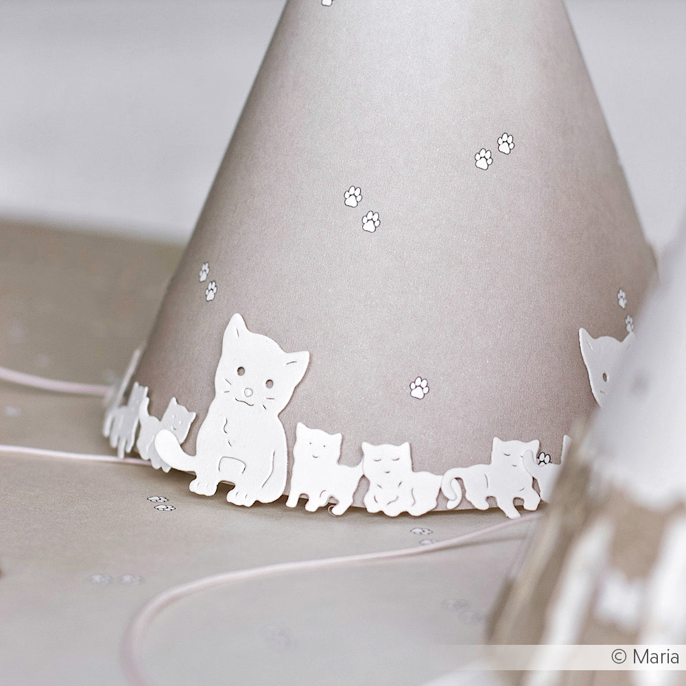 Ein Partyhut mit weißen Katzenmotiven und Pfotenabdrücken auf hellem Grund, inspiriert von der Alexandra Renke Motivstanze Kätzchen Familie und hergestellt mit hochwertigen Stahlstempeln.