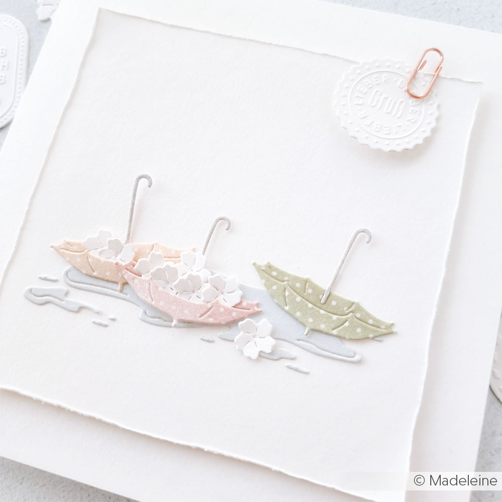 Drei pastellfarbene Regenschirme mit kleinen Blumen und Pfützen schmücken eine strukturierte weiße Karte, die mit einem Papiersiegel veredelt wurde. Diese Karte wurde mit der Motivstanze Hundewetter von Alexandra Renke hergestellt und zeigt die Vielseitigkeit von Stanzen für Designpapiere für handgemachte Karten.