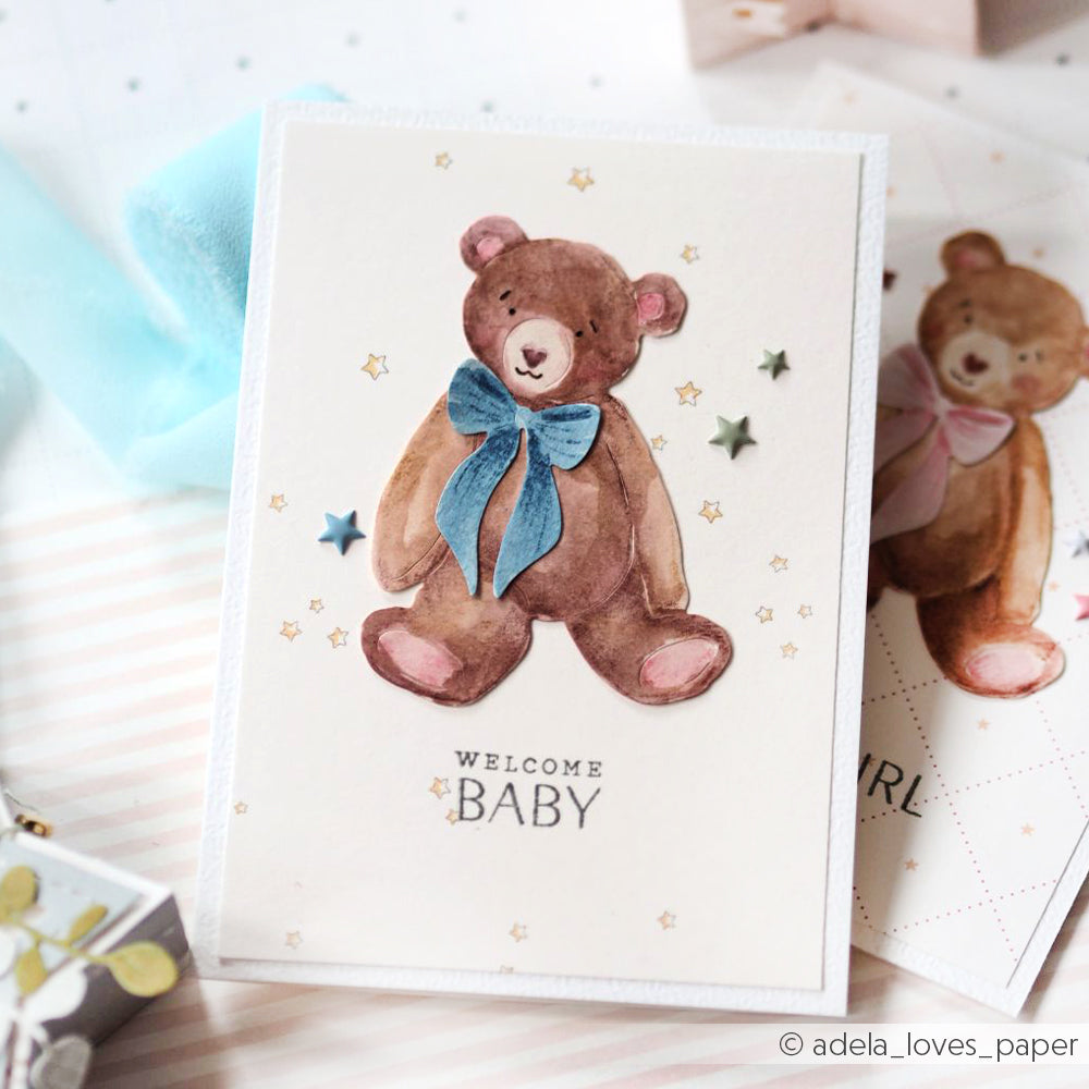 Die Grußkarte von Alexandra Renke zeigt einen aquarellierten Teddybären mit einer blauen Schleife, Sternen und "WELCOME BABY" auf der Vorderseite - gefertigt mit hochwertigen Designpapieren für einen charmanten Touch.