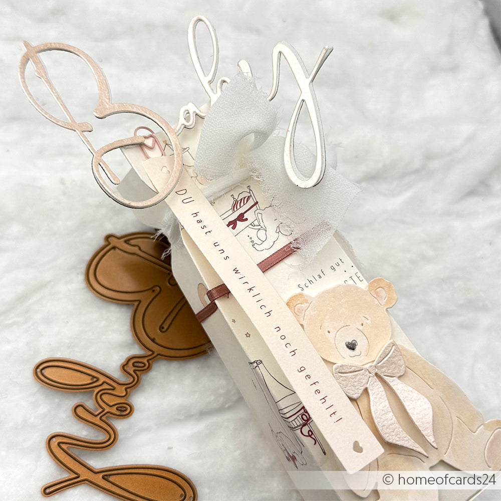Dekoratives Geschenk von Alexandra Renke mit "Baby"-Text, Anhängern und einem niedlichen Bären, der mit DieCut Systemen auf einem weichen weißen Hintergrund ausgestanzt wurde.