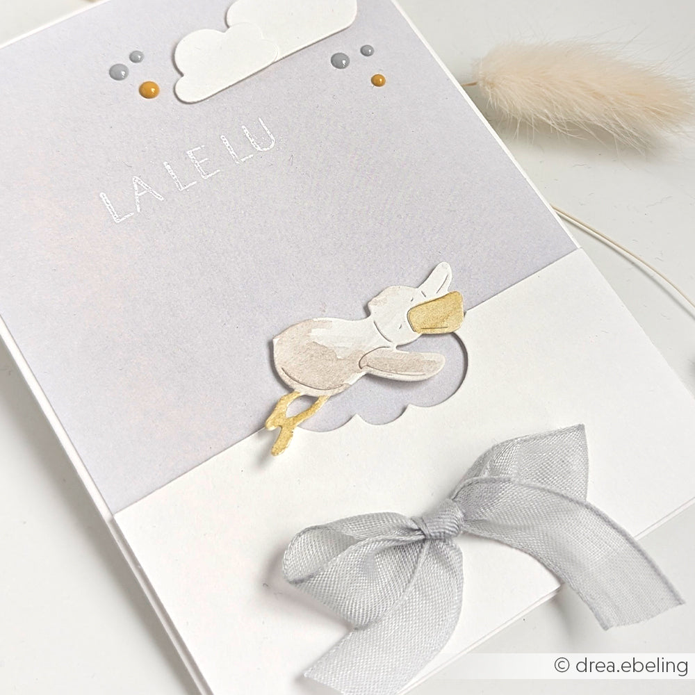 Grußkarte von Alexandra Renke mit fliegendem Storch, Wolken, Punkten, silberner Schleife und dem Text LA LE LU auf Weiß - ideal zur Kombination mit Designpapieren oder DieCut Systemen für kreative Projekte.