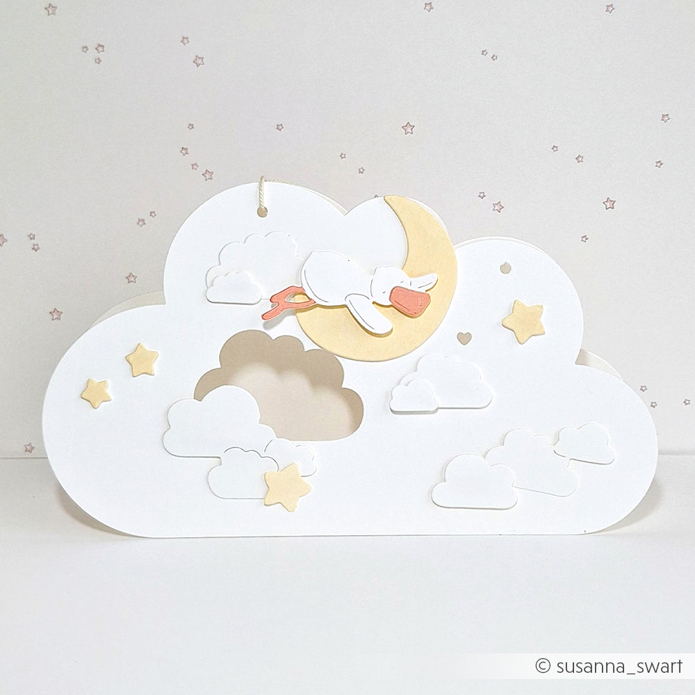 Papierkunstwerk eines schlafenden Storches auf einer Mondsichel, umgeben von Wolken und Sternen, hergestellt mit Alexandra Renke DieCut Systemen für präzise Details auf einem wolkigen weißen Hintergrund.