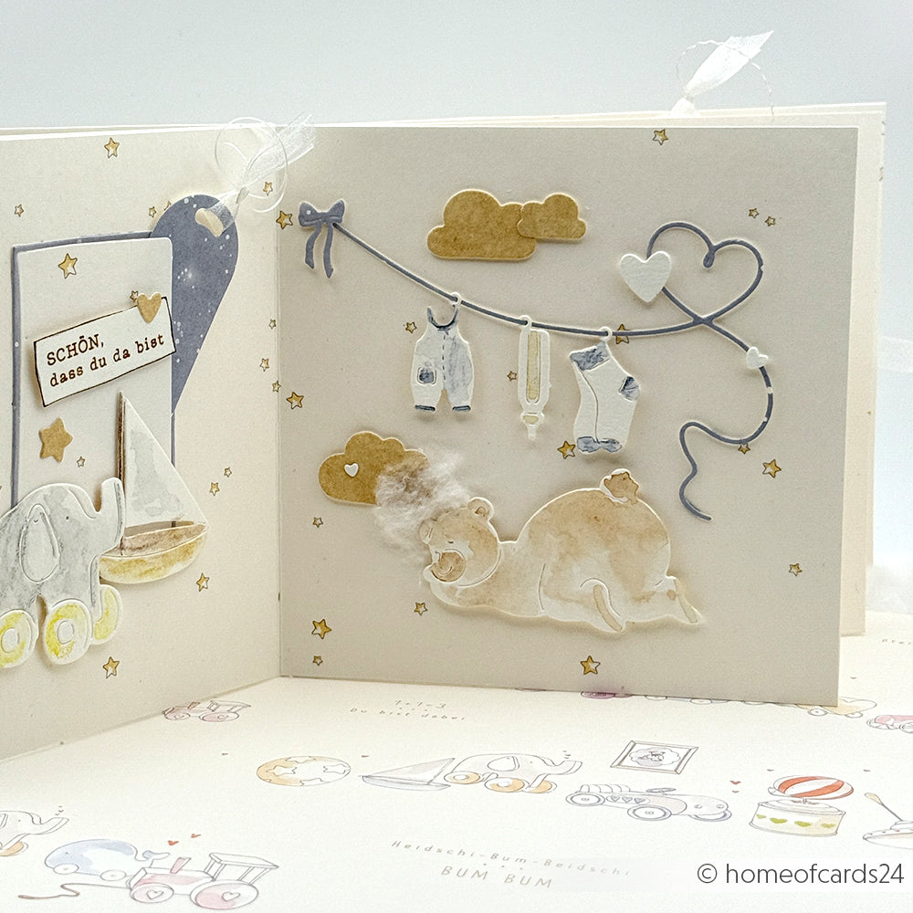 Handgemachte Babykarte von Alexandra Renke, hergestellt mit Motivstanze und Designpapier, mit einem Bären, einem Elefanten, Babykleidung auf einer Leine und goldenen Sternen auf weißem Hintergrund.