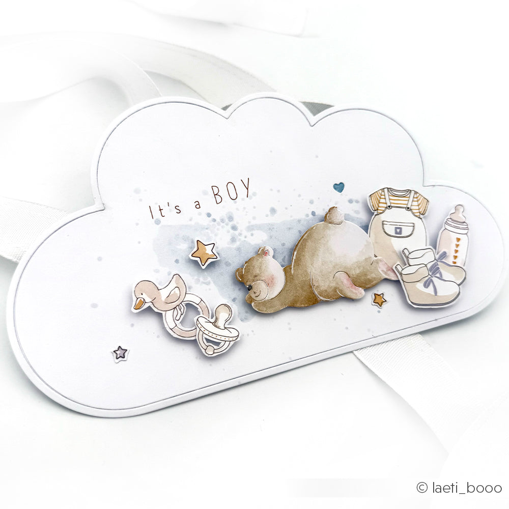 Die wolkenförmige Karte von Alexandra Renke enthält den Text "It's a BOY", DieCut-Systeme-Details und niedliche Illustrationen eines Teddybären, eines Schnullers, von Babykleidung und eines Fläschchens - perfekt für die Begrüßung Ihres Kleinen.