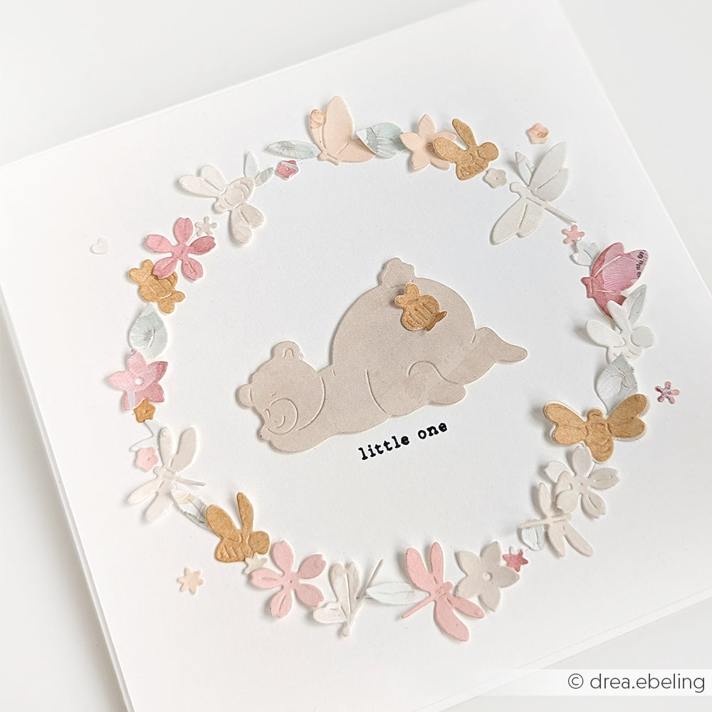 Eine Karte von Alexandra Renke mit einem Bären und einem Baby in einem pastellfarbenen Blumen- und Schmetterlingskranz, gestaltet mit zarten Designpapieren.