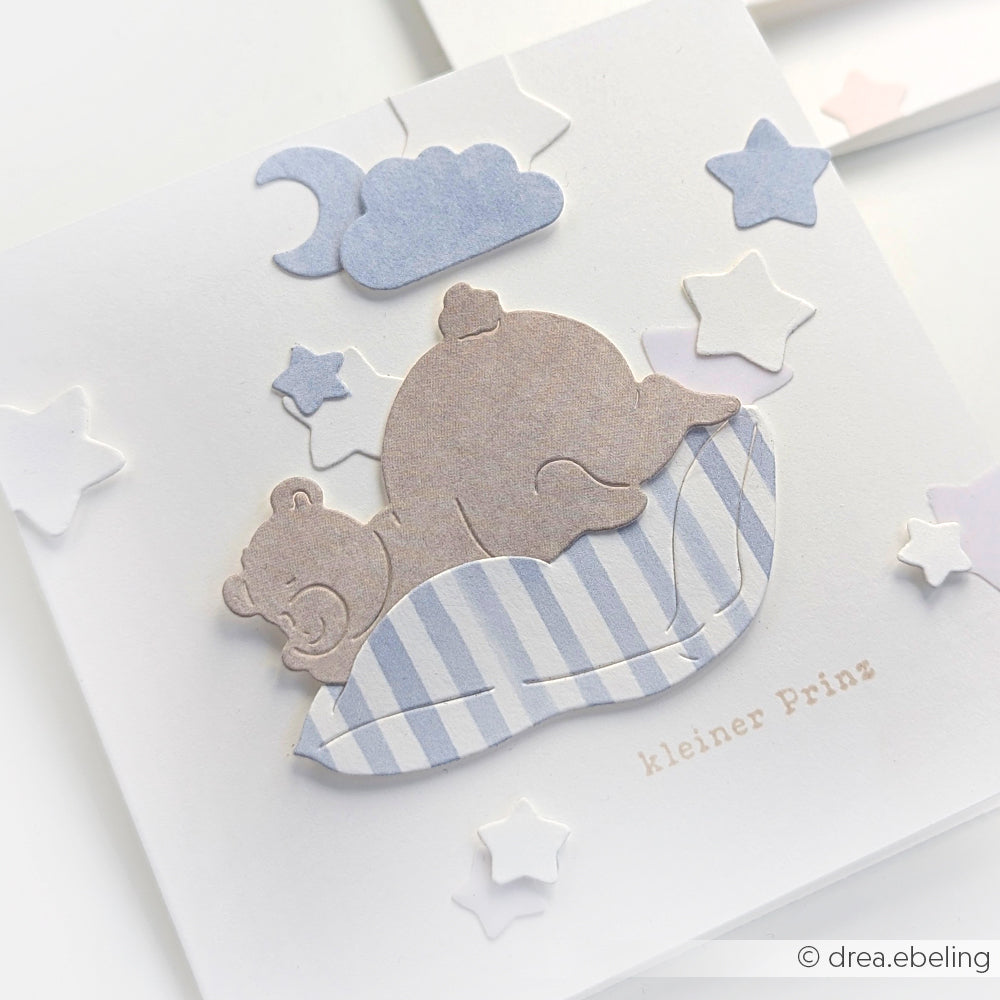 Papierausschnitt eines schlafenden Bären auf einem gestreiften Kissen mit Sternen, Wolken und dem Text "Kleiner Prinz" - hergestellt mit Alexandra Renke DieCut Systemen und charmanten Designpapieren für skurrilen Charme.