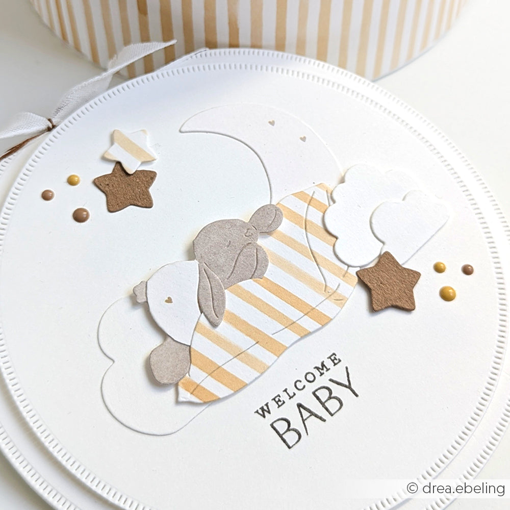 Runde Karte mit Stanzformen von Alexandra Renke, die einen schlafenden Teddybären auf Wolken und Sternen zeigt, mit dem Text "WELCOME BABY" am unteren Rand.