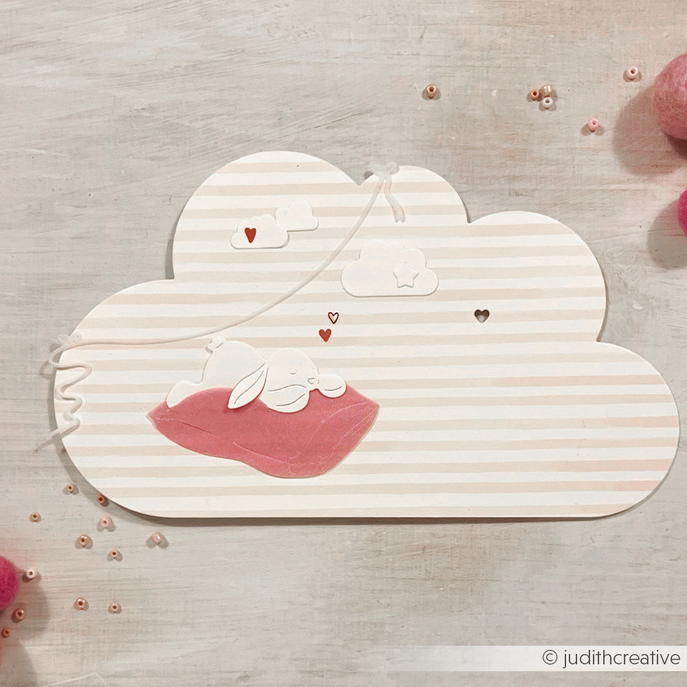 Ein verschlafenes Häschen ruht auf einer rosa Decke auf einer gestreiften Wolke, umgeben von kleinen Herzen - ideal zum Basteln mit der Motivstanze Müdes Häschen von Alexandra Renke oder zum Verzieren von Projekten mit Alexandra Renke Stanzen für Designpapier.