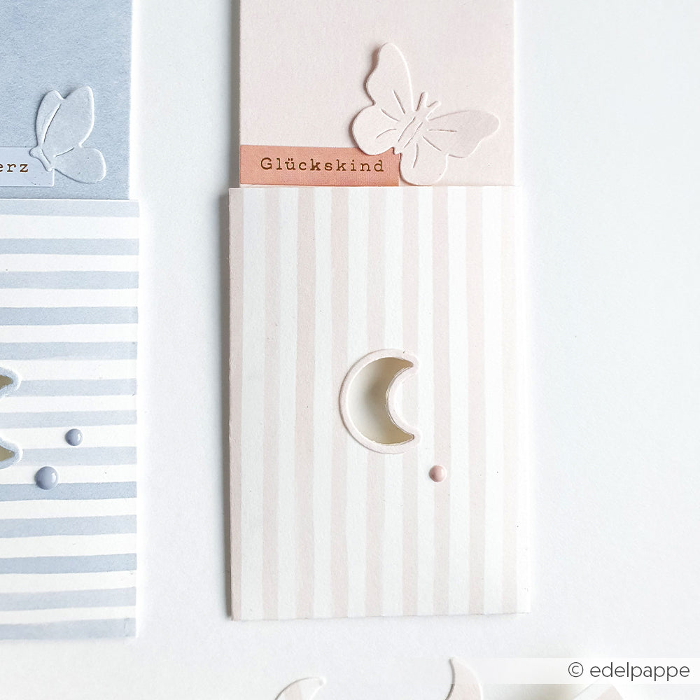 Eine pastellfarbene, gestreifte Karte mit Schmetterlingsausschnitt, Mondsichelfenster, Glückskind-Etikett und zarten Designpapieren - alles mit Produkten von Alexandra Renke hergestellt.
