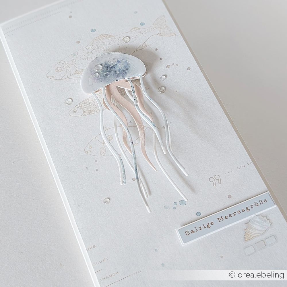 Handgefertigte Karte mit einer 3D-Qualle, die mit der Alexandra Renke Motivstanze „Qualle“ gestaltet wurde, zarten Fischskizzen und dem Text „Salzige Meeresgrüße“ auf weißem Hintergrund. Hergestellt aus hochwertigem Alexandra Renke Designpapier (200 g) und mit Details aus den Alexandra Renke Stanzen aus Stahl verziert, die für zusätzliche Tiefe sorgen.