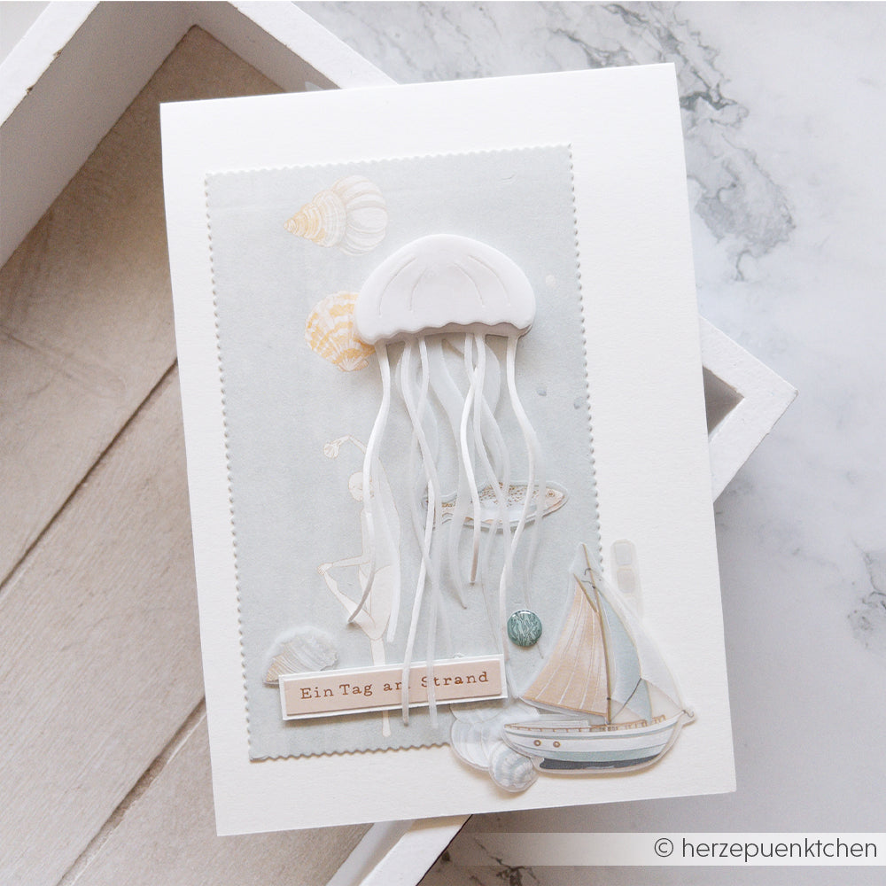 Handgefertigte Karte aus Designpapieren von Alexandra Renke mit einer Qualle, die mit der Motivstanze „Qualle“ ausgestanzt wurde, Muscheln, einem Segelboot und dem Schriftzug „Ein Tag am Strand“ auf weißem Hintergrund.
