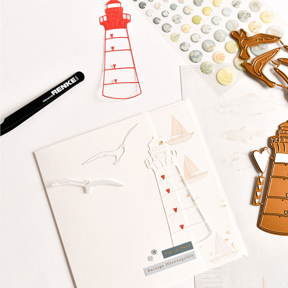 Papierbastelmaterialien von Alexandra Renke mit Leuchtturm-Motiv, Designpapiere, Metallstanzformen aus dem Motivstanze-Set „Möwen“, Aufkleber, eine Pinzette und eine Grußkarte.