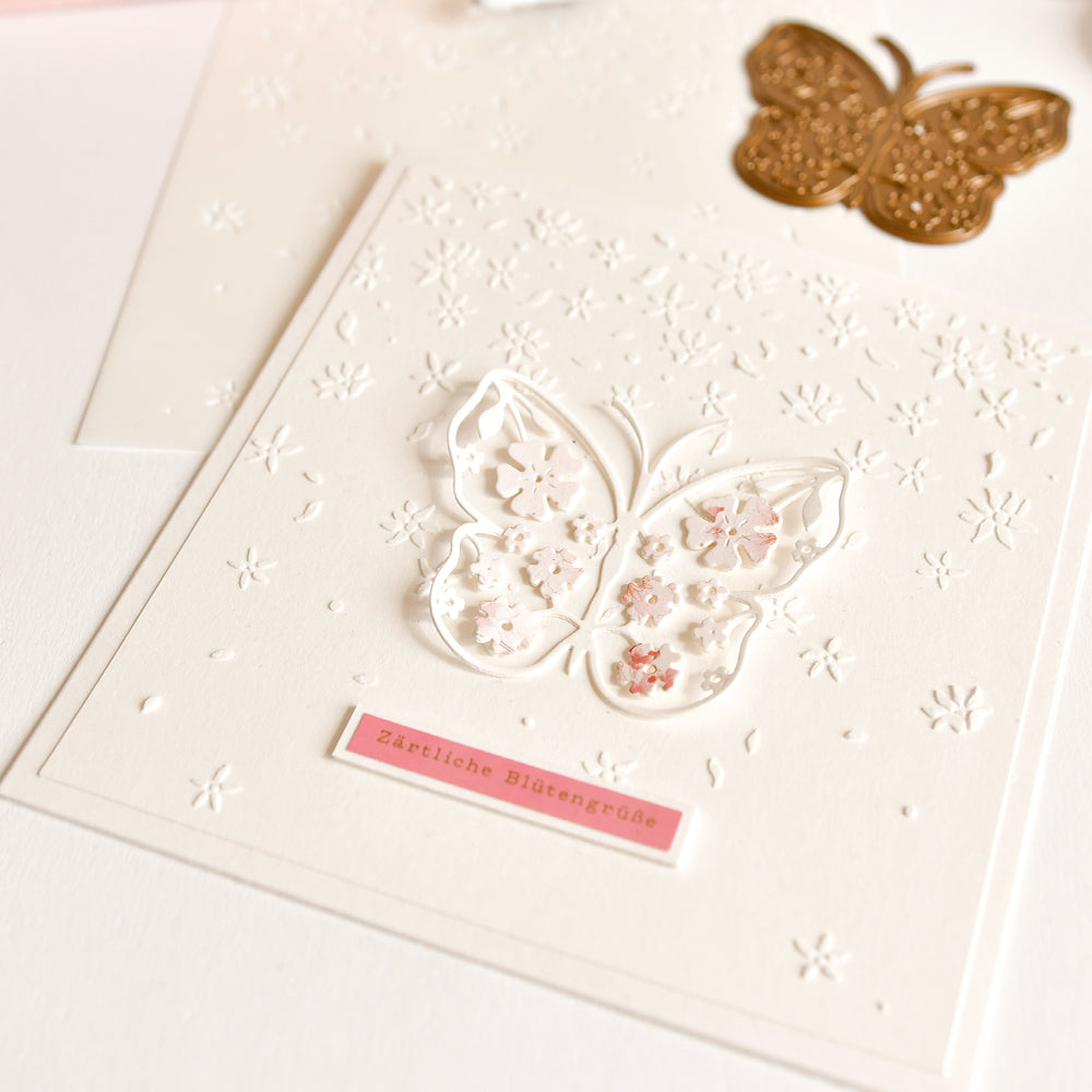 Weiße Karte mit einem filigranen Schmetterlingsausschnitt, gestanzt mit der Motivstanze „Blumiger Schmetterling“ von Alexandra Renke, floraler Prägung auf hochwertigem Designpapier und einem rosa Etikett mit der Aufschrift „Zärtliche Blütengrüße“.