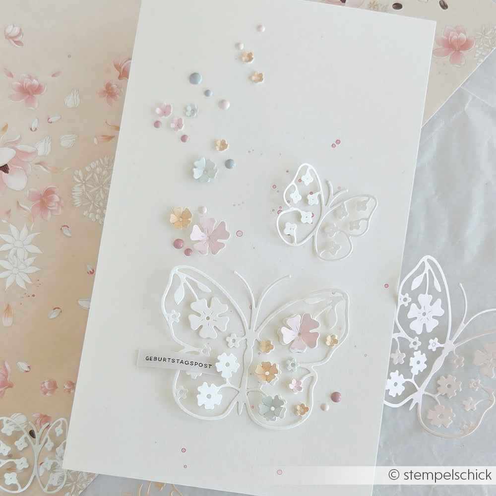 Weiße Grußkarte von Alexandra Renke mit zarten Schmetterlings- und Blumenausschnitten, sanften Pastell-Akzenten aus Designpapier und einem „Geburtstagspost“-Anhänger.