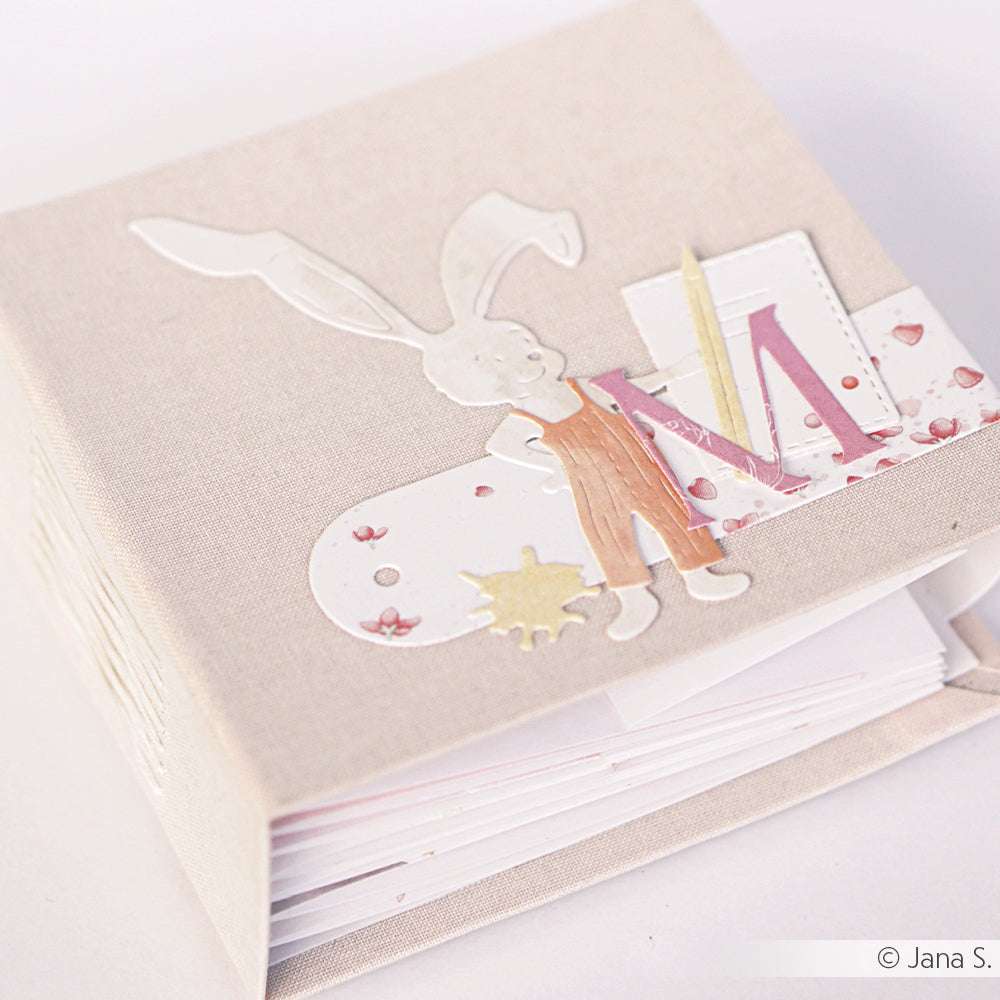 Handgemachtes Scrapbook von Alexandra Renke mit einem Hasen, dem Buchstaben M aus der Motivstanze Alphabet Dezember und dekorativen Akzenten auf einem beigen Stoffeinband.