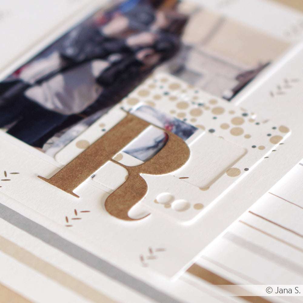 Scrapbook-Seite mit einem großen goldenen Buchstaben R, hergestellt mit der Alexandra Renke Motivstanze Alphabet Groß, überlagert auf Designpapier mit einem unscharfen Foto im Hintergrund.