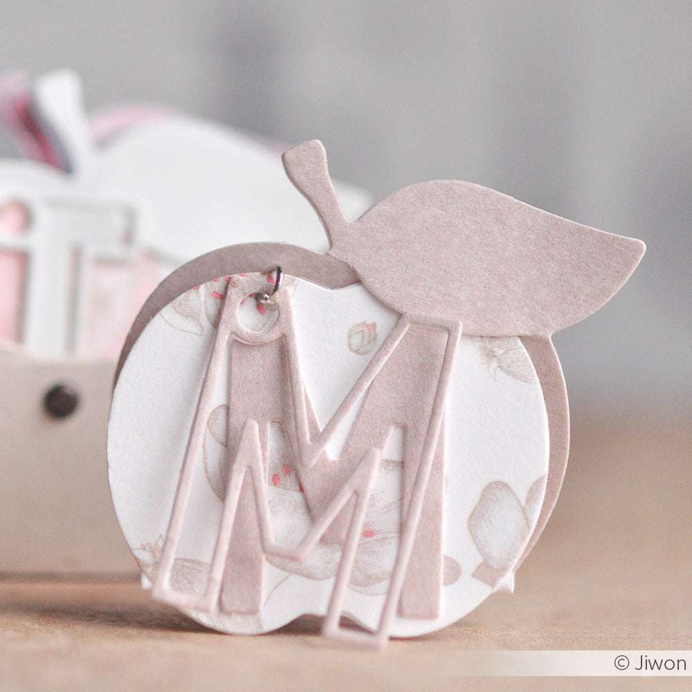 Ein pastellrosa apfelförmiger Anhänger mit einem großen ausgeschnittenen Buchstaben M, gefertigt aus Alexandra Renke Designpapieren und Motivstanze Konturbuchstaben, ruht auf einer hellen Oberfläche.
