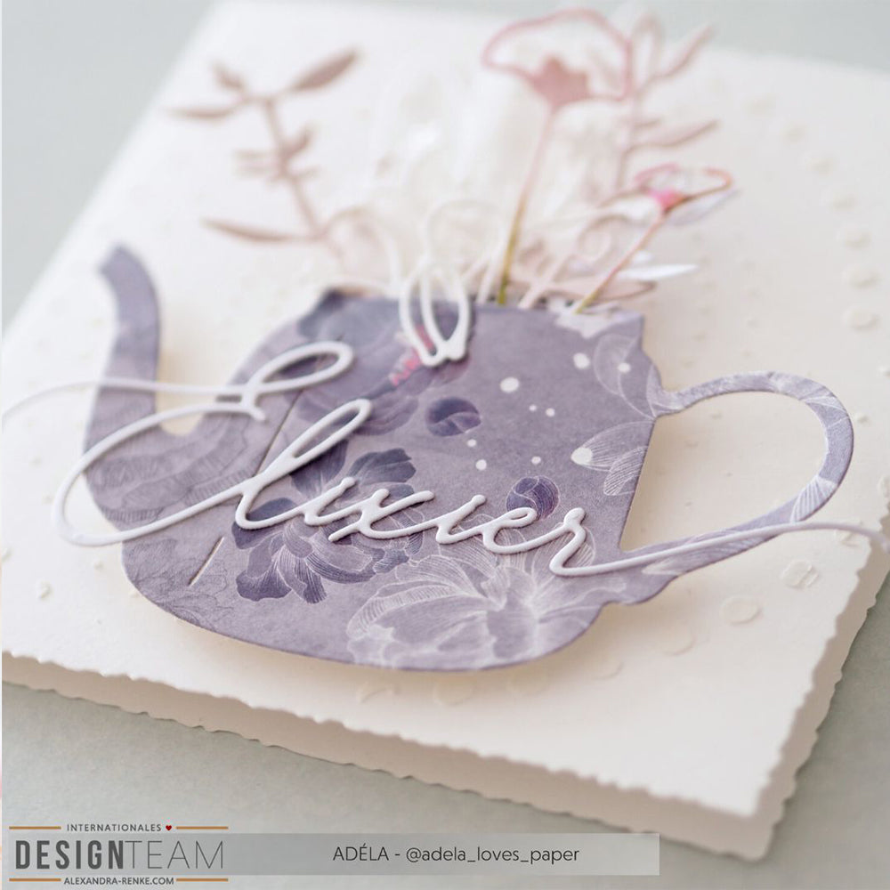 Handgefertigte Karte von Alexandra Renke mit einer lilafarbenen Teekanne mit Blumen, weißem "Elixier"-Schriftzug und zarten Papierblumen, die mit der Motivstanze auf hochwertigem Designpapier hergestellt wurden, vor einem strahlend weißen Hintergrund.