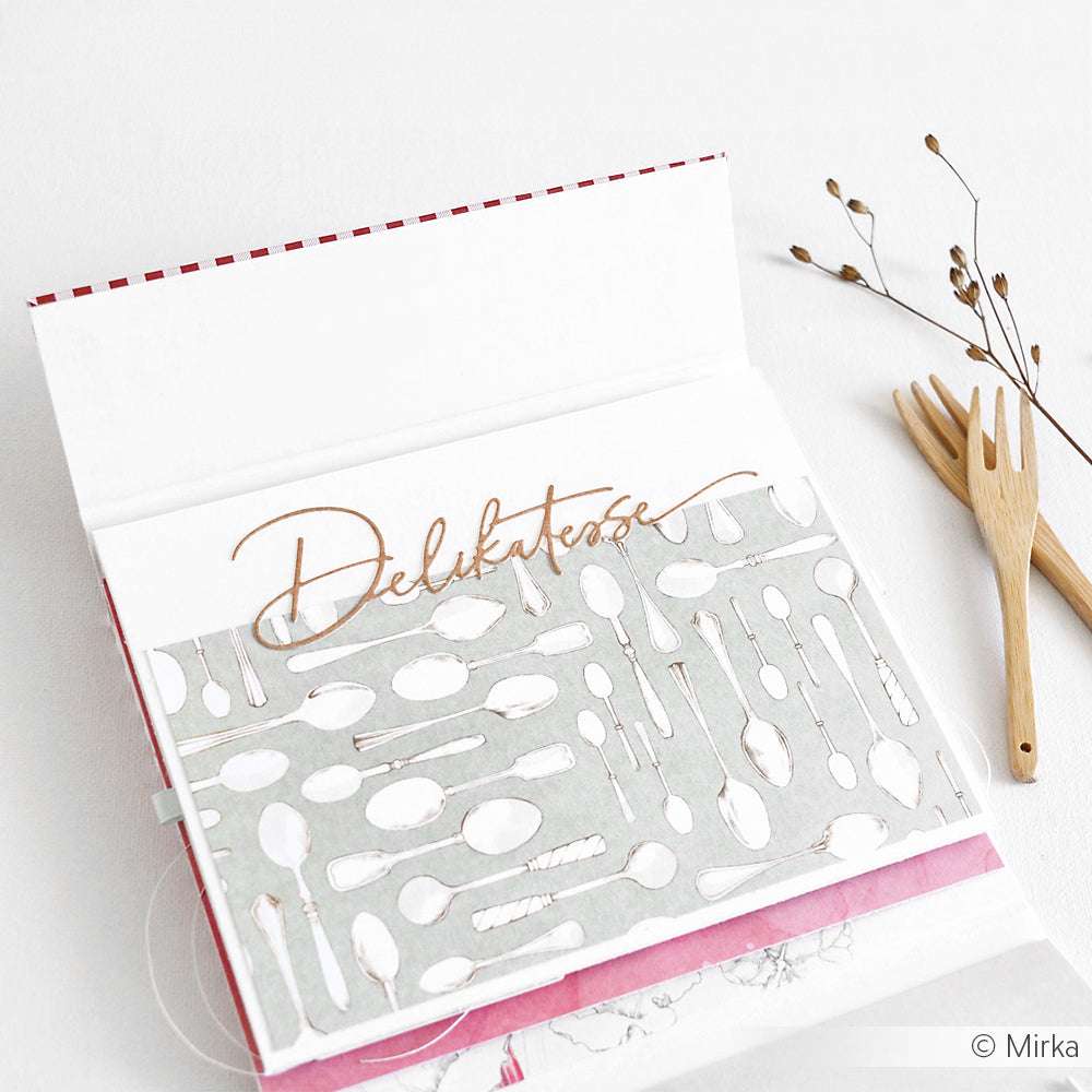 Aufgeschlagenes Buch mit "Delikatesse" im Inneren, einer Holzgabel und getrockneten Pflanzen auf weißem Grund - eine ideale Inspiration für Alexandra Renke Designpapiere oder kreative Motivstanze-Projekte.
