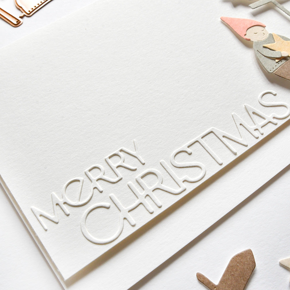 Weiße Karte von Alexandra Renke mit "Frohe Weihnachten" in erhabenen Buchstaben, mit DieCut Systeme Papierausschnitten eines Gnoms und festlichen Formen.