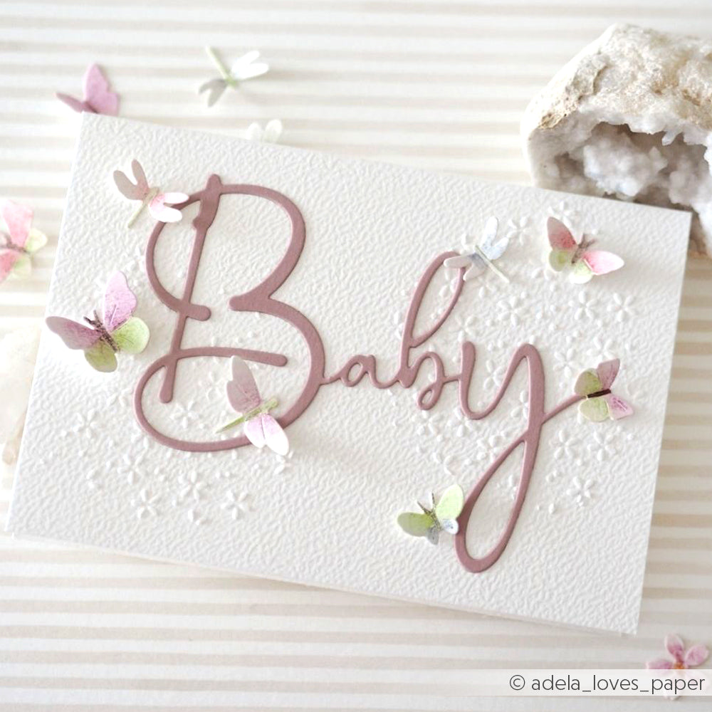 Eine weiße Karte mit dem Schriftzug "Baby" in rosa Schrift, gestaltet mit der Alexandra Renke Motivstanze Baby und pastellfarbenen Schmetterlingen von Alexandra Renke Designpapiere auf gestreiftem Hintergrund.