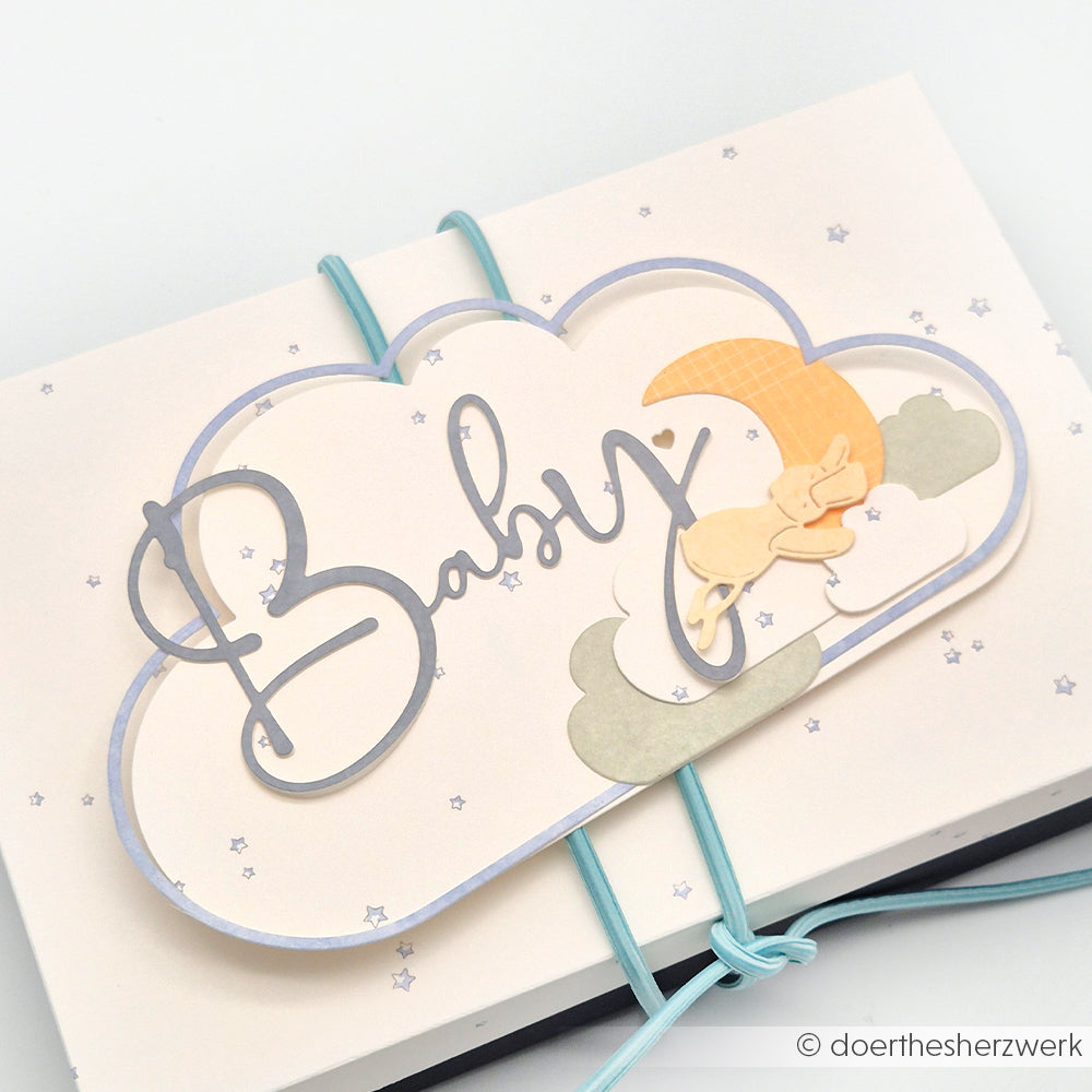 Wolkenförmige Karte von Alexandra Renke, hergestellt mit DieCut Systemen, mit dem Text "Baby", Sternen, einem Mond und einem schlafenden Häschen, gebunden mit blauen Gummibändern.
