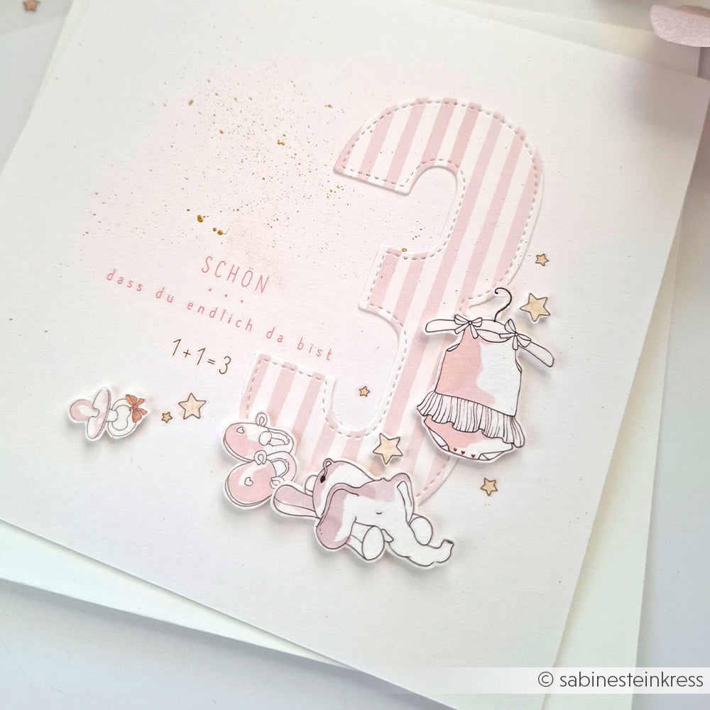 Rosa-weiße Karte mit einer großen Zahl 3, hergestellt mit Alexandra Renke Motivstanze Zahlen Dies, verziert mit Babykleidung, Schuhen, Sternen und deutschem Text.