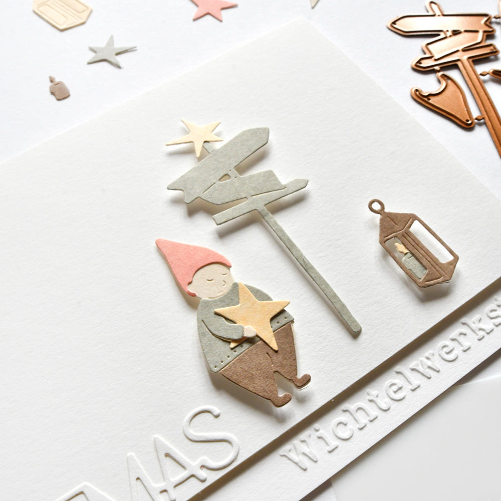 Ein mit Alexandra Renke DieCut Systeme gestalteter Papierzwerg steht neben einem Wegweiser und einer Laterne und hält einen Stern auf einer weißen Karte mit geprägtem Text.