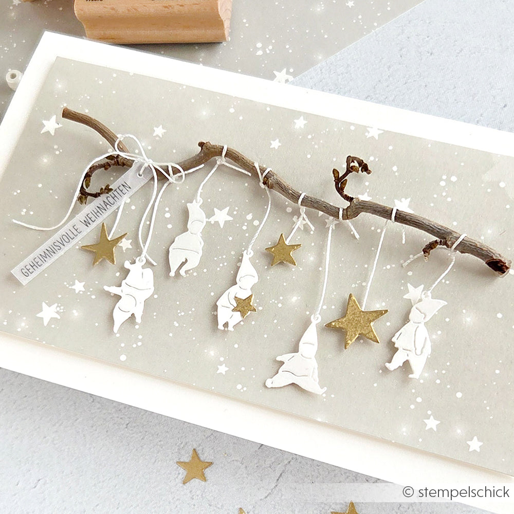 Eine Karte von Alexandra Renke mit DieCut Systemen mit einem Zweig, weißem Figurenschmuck, hängenden Goldsternen und einem deutschen Weihnachtsanhänger.