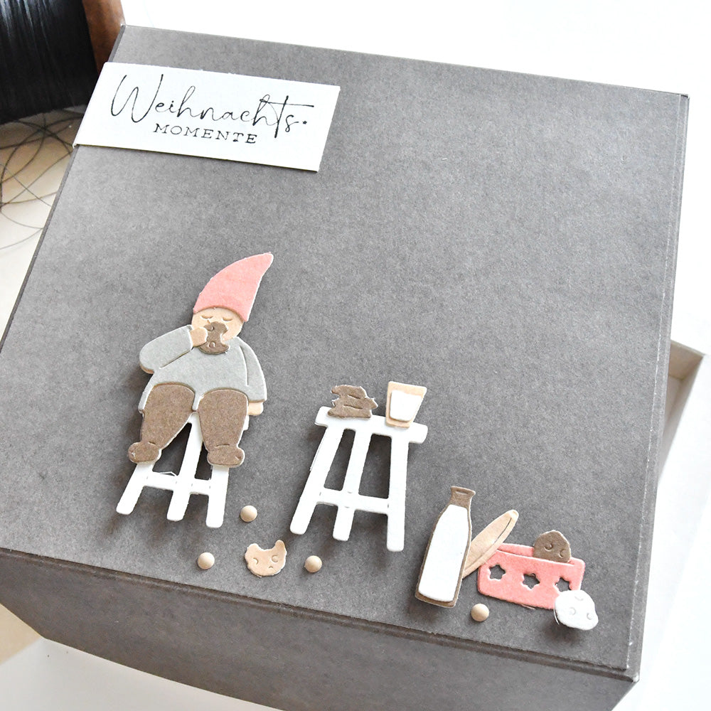 Eine graue Box von Alexandra Renke, geschmückt mit einem Wichtel, Hockern, Knabbereien und einem "Weihnachts Momente"-Anhänger, enthält die bezaubernde Motivstanze Wichtel Nascherei und Stanzsysteme zum festlichen Basteln.