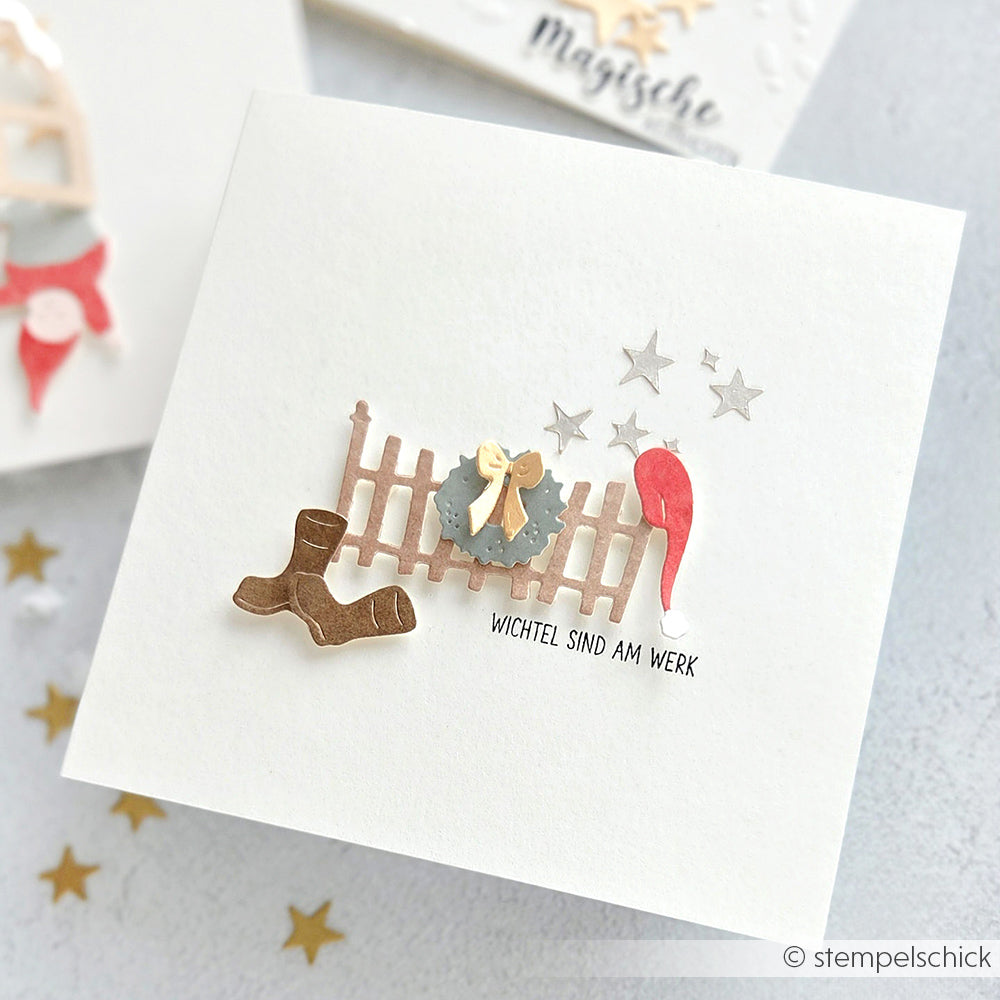 Handgefertigte Karte von Alexandra Renke mit einem Wichtel auf einer Motivstanze Kleiner Zaun mit Kranz, roter Mütze, Stiefeln, silbernen Sternen, Text "Wichtel sind am Werk", hergestellt aus hochwertigem 200g Designpapier.
