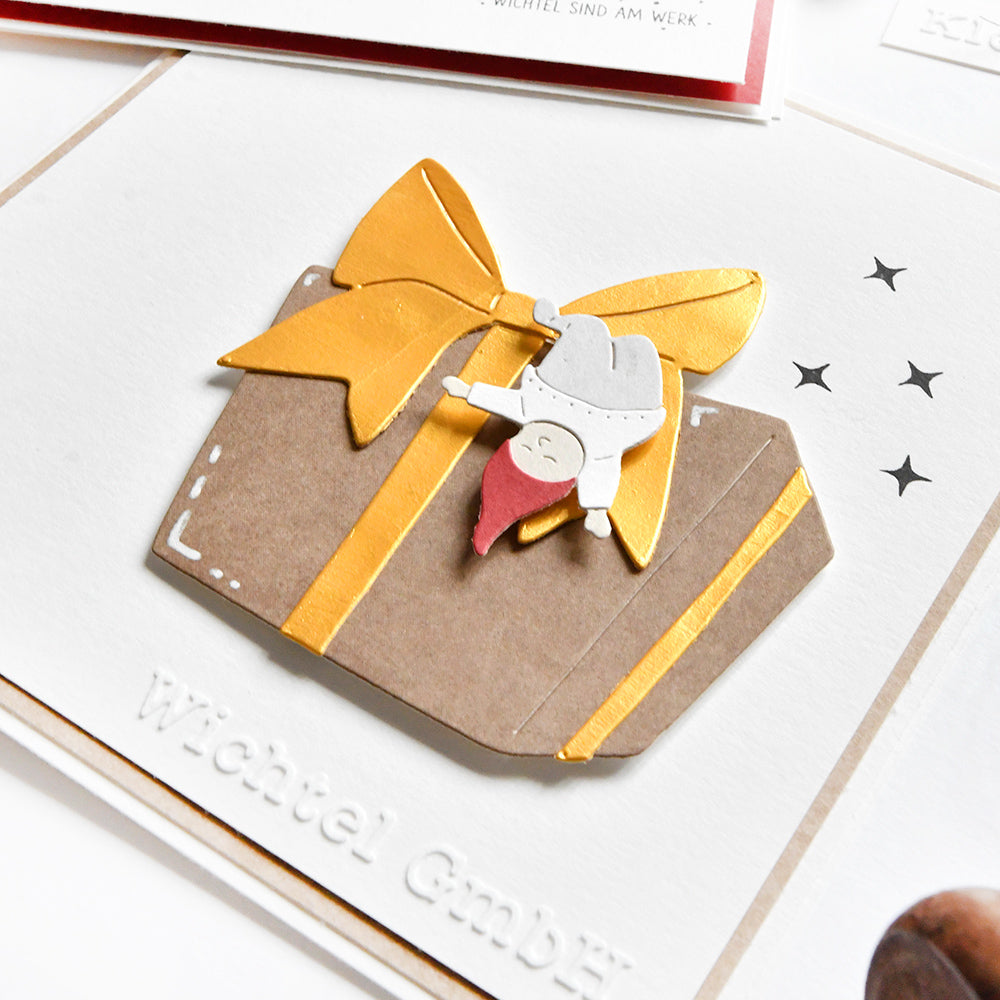 Eine handgefertigte Karte von Alexandra Renke mit einer großen Geschenkschachtel mit goldener Schleife und einem kleinen Gnom auf der Spitze, hergestellt mit Motivstanze Wichtel für zusätzlichen Charme.